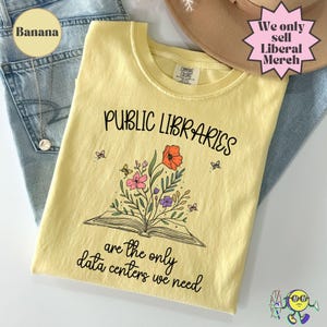 May include: A pale yellow crew neck t-shirt featuring a black graphic of an open book with colorful flowers and butterflies emerging from the pages. The text reads Public Libraries are the only data centers we need in a black cursive and print font.