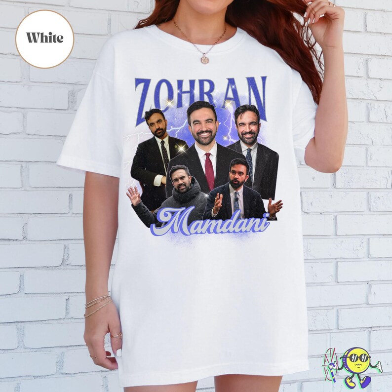 White oversized T-shirt featuring multiple images of a man in different poses and outfits, mainly in suits and a hoodie. The top text reads "ZOHRAN" in bold blue stylized letters with sparkles and lightning effects in the background. Below the images, "Mamdani" is written in a cursive blue font with a glowing effect.