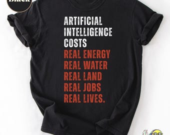 Puede incluir: Una camiseta negra de manga corta con texto blanco que dice ARTIFICIAL INTELLIGENCE COSTS y texto rojo que dice REAL ENERGY, REAL WATER, REAL LAND, REAL JOBS, REAL LIVES. Una pequeña insignia circular en la esquina superior izquierda dice Black.
