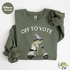 May include: A military green crewneck sweatshirt featuring the white text OFF TO VOTE. The graphic depicts a green frog wearing a light purple pointed hat, riding a light gray motor scooter while holding a white ballot paper and wearing a voting pin.