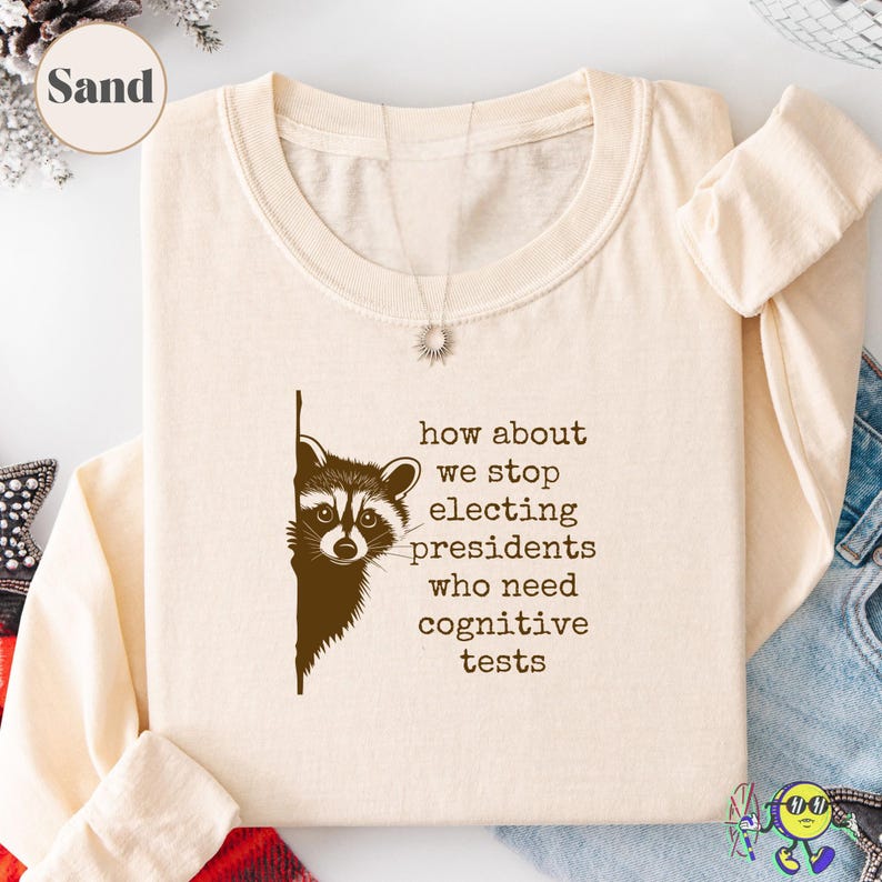 Sand-colored long-sleeve shirt with a round neck, featuring a brown graphic of a raccoon peeking from behind a vertical line. Next to the raccoon, text reads: "how about we stop electing presidents who need cognitive tests" in typewriter-style font. The shirt is neatly folded with one sleeve rolled. A silver sunburst necklace lays on top near the collar. Denim and plaid fabric partially visible around the shirt.