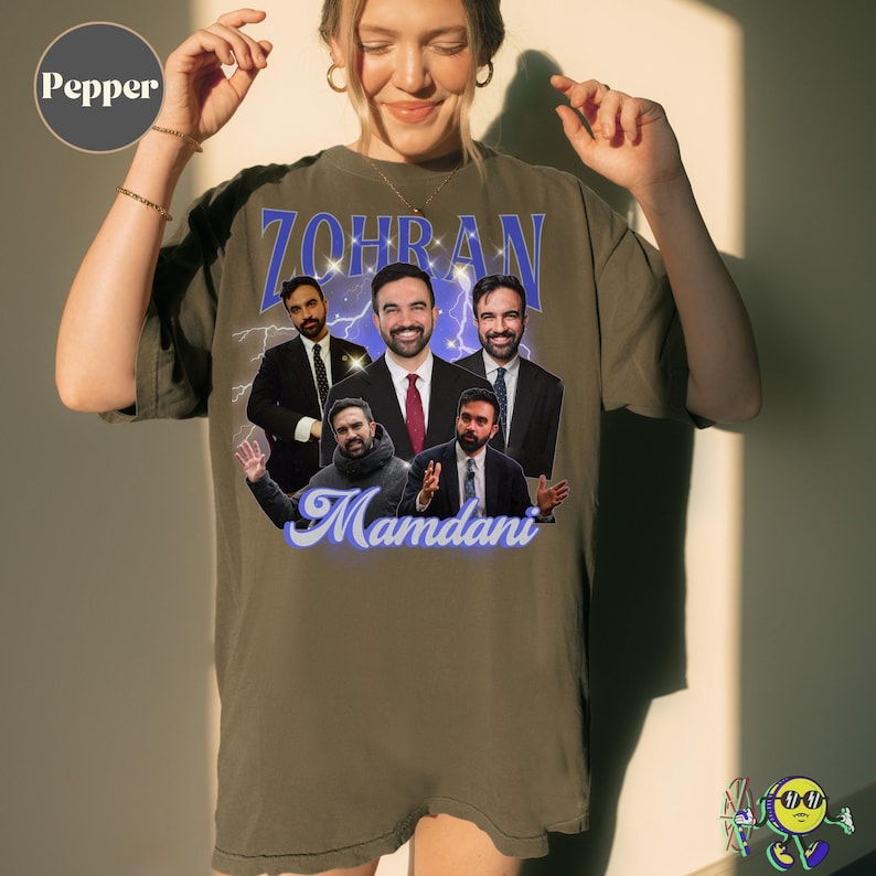 A model wearing an oversized Pepper-colored t-shirt featuring a collage of five photos of a man in different poses and suits. Above the images is large blue text "ZOHRAN" with lightning effects, below in blue script is the name "Mamdani." The shirt has a casual fit with rolled sleeves.