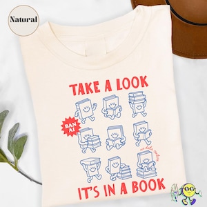 May include: A natural-colored t-shirt featuring red text that reads "TAKE A LOOK" and "IT'S IN A BOOK". The graphic includes blue line-art illustrations of smiling books in various poses, along with a red starburst icon labeled "BAN AI".