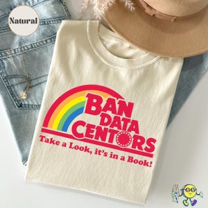 Puede incluir: Una camiseta de color crema con un gráfico de arcoíris y texto rojo que dice BAN DATA CENTORS y Take a Look, it's in a Book! La camiseta se muestra junto a tela de mezclilla azul, un sombrero beige de ala ancha y un collar dorado fino.