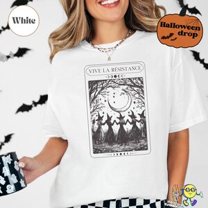 White t-shirt worn by a model, featuring a black vintage tarot card-style design of five witches holding hands beneath a crescent moon and stars, framed by leafless trees. Above the illustration, “VIVE LA RÉSISTANCE” is written. The print includes small crescent moon phases along the top and bottom edges of the card. The model holds a black mug with white ghost faces. A small orange pumpkin graphic says “Halloween drop.”