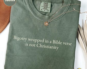 Può includere: Una maglietta verde muschio con la scritta bianca "Bigotry wrapped in a Bible verse is not Christianity". Una collana d'oro con un ciondolo è appoggiata sulla maglietta. La maglietta ha un'etichetta Comfort Colors.