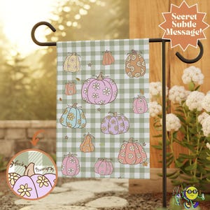 May include: A garden flag featuring a pattern of pumpkins in various colors, including pink, orange, and blue, adorned with floral designs. The pumpkins are set against a green and white checkered background. The flag is displayed on a black metal stand.