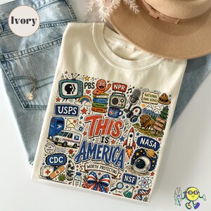 May include: An ivory t-shirt featuring a colourful graphic print with the text "This is America Worth Protecting" surrounded by illustrations of icons including a PBS television, a USPS truck, a rocket, a NASA logo, a bison, and scientific symbols.