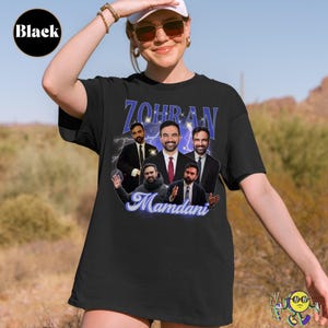 Black t-shirt featuring a collage of six images of a man in a suit with various expressions and gestures. Above the images is the name "Zohran" in large blue letters with sparkles and a lightning background. Below, the name "Mamdani" is written in a flowing, light blue script. A model wears the shirt outdoors, with a desert landscape behind.