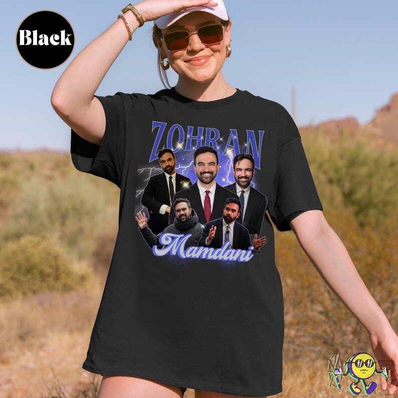 Black t-shirt featuring a collage of six images of a man in a suit with various expressions and gestures. Above the images is the name "Zohran" in large blue letters with sparkles and a lightning background. Below, the name "Mamdani" is written in a flowing, light blue script. A model wears the shirt outdoors, with a desert landscape behind.