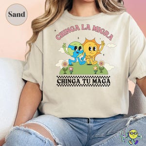May include: Sand-colored t-shirt featuring a cartoon Earth and Sun character holding hands, with the text "CHINGA LA MIGRA" and "CHINGA TU MAGA" in pink and black. The design includes flowers and a checkered border.