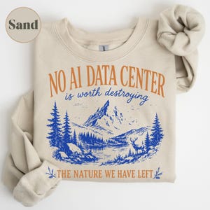 Może przedstawiać: Bluza z okrągłym dekoltem w kolorze piaskowym z nadrukiem graficznym w kolorze pomarańczowym i niebieskim. Tekst brzmi NO AI DATA CENTER is worth destroying THE NATURE WE HAVE LEFT. Ilustracja przedstawia górski krajobraz z sosnami i jeleniem.