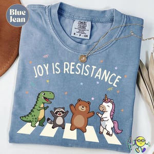 Może przedstawiać: Błękitna koszulka z napisem "JOY IS RESISTANCE" i grafiką przedstawiającą dinozaura, szopa pracza, niedźwiedzia i jednorożca przechodzących przez przejście dla pieszych. Koszulka ma metkę Comfort Colors i złoty naszyjnik.