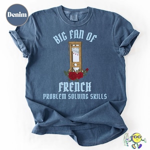 May include: Denim blue t-shirt with the text "BIG FAN OF FRENCH PROBLEM SOLVING SKILLS". The shirt features a graphic of a guillotine with red roses. The shirt is a crew neck and has rolled-up sleeves.