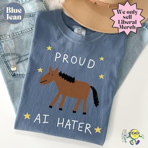 Pode incluir: Uma t-shirt azul com uma ilustração de um cavalo castanho com crina e cauda pretas. O texto PROUD AI HATER está impresso a branco, rodeado por estrelas amarelas. A t-shirt tem uma etiqueta Comfort Colors no decote interior.