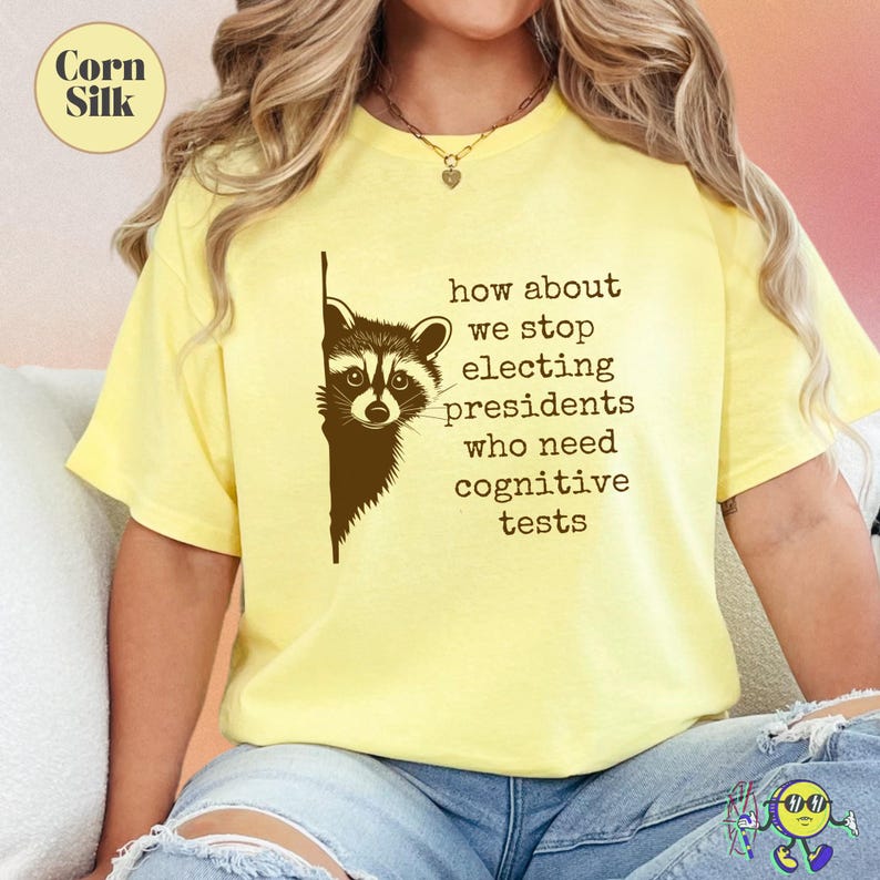 Yellow "Corn Silk" t-shirt worn by a model, featuring a dark brown graphic of a raccoon peeking from the left side, next to typewriter-style text reading, "how about we stop electing presidents who need cognitive tests." The shirt has a relaxed fit and is paired with light blue ripped denim shorts.