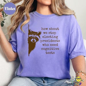 A violet t-shirt worn by a model features a brown raccoon peeking from the left side. To the right of the raccoon, brown typewriter-style text reads: "how about we stop electing presidents who need cognitive tests." The shirt has a casual fit with rolled short sleeves.