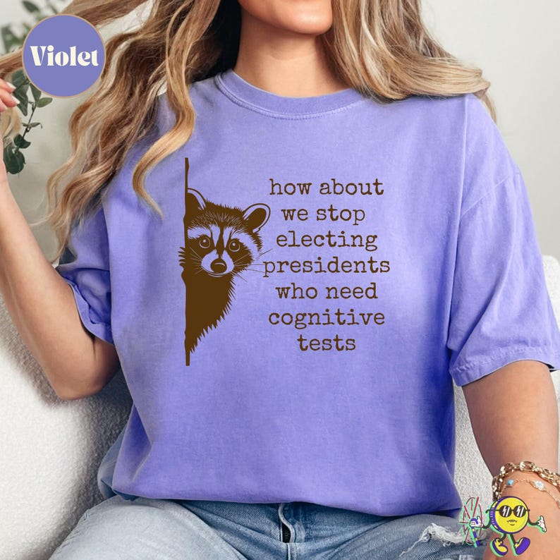 A violet t-shirt worn by a model features a brown raccoon peeking from the left side. To the right of the raccoon, brown typewriter-style text reads: "how about we stop electing presidents who need cognitive tests." The shirt has a casual fit with rolled short sleeves.