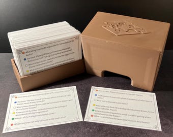 May include: A tan plastic card box with a lid featuring a geometric emblem, containing a stack of white trivia cards. Two additional white cards with printed questions and colored bullet points are placed in front of the box on a dark surface.