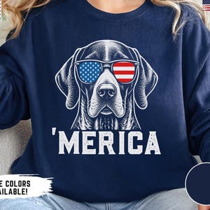 May include: A navy blue long-sleeve sweatshirt featuring a white graphic illustration of a dog wearing sunglasses with an American flag pattern. Below the dog, the text reads 'MERICA in white capital letters. The design is centred on the chest.