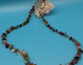 May include: A necklace made of small, irregularly shaped, colorful gemstones. The stones are strung together with a silver clasp. The necklace is displayed against a light blue background, with a white crystal formation behind the clasp.