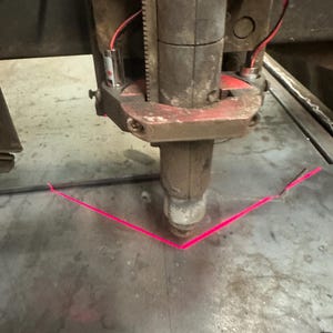 May include: A close-up of a metal cutting machine with a red laser beam pointing down at a metal surface. The laser beam is creating a V-shaped pattern on the metal.