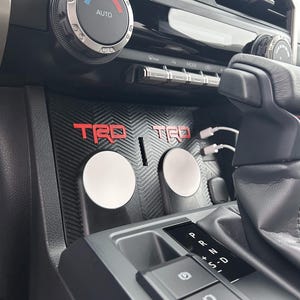 May include: Black car interior with a climate control dial and buttons. A center console features the red TRD logo and two silver-colored circular objects. USB cables are visible.