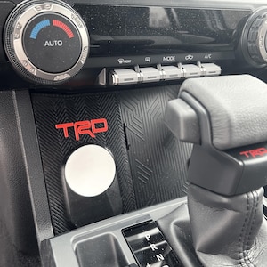 May include: Interior car console with a black textured surface and red "TRD" logo. Features include climate control dials, mode buttons, and a gear shift lever. A white circular object is also visible.