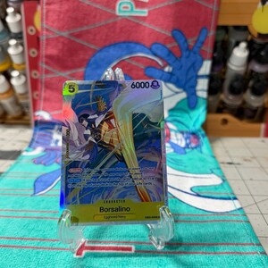 May include: A holographic One Piece trading card featuring the character Borsalino. The card displays a cost of 5, a power of 6000, and the text Borsalino, Egghead/Navy. It is held in a clear plastic stand, showcasing vibrant yellow and blue artwork.