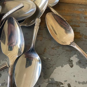 May include: A pile of silver spoons, some with ornate handles, are stacked on a rustic wooden surface. The spoons are shiny and reflect light.