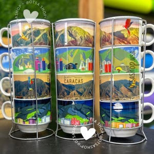 May include: Set of four white ceramic mugs stacked in a metal stand. Each mug features a different scenic image of Caracas, Venezuela. The mugs are decorated with colorful illustrations of mountains, buildings, and a parrot.