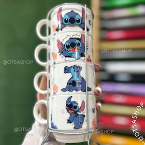 Może przedstawiać: Zestaw czterech białych ceramicznych kubków z postacią Stitch z Lilo & Stitch. Każdy kubek ma inny wzór Stitch, z niebieskimi i różowymi akcentami. Kubki są trzymane w srebrnym metalowym stojaku.