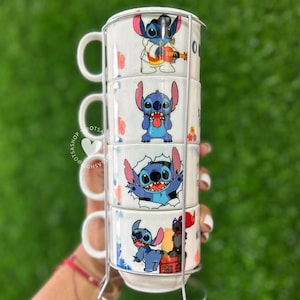 Może przedstawiać: Zestaw kubków do układania w stosy z postacią Stitcha z Lilo & Stitch. Każdy biały ceramiczny kubek ma inny wzór Stitcha, ze metalowym stojakiem. Kubki są ułożone w stosy pionowo, trzymane przez srebrny metalowy stojak i mają białe uchwyty.