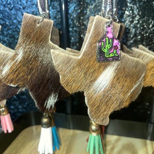 May include: A set of brown and white cowhide Texas-shaped keychains with a pink and green cactus charm. Each keychain has a tassel with a different color: pink, white, green, and red.