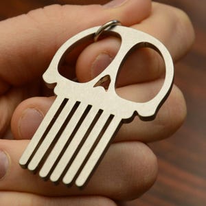 May include: A silver skull-shaped keychain with a comb-like design on the bottom. The keychain is held in a hand.