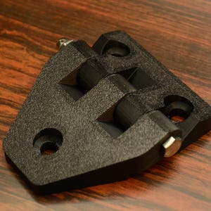 May include: A black, textured hinge with two pivoting sections and silver-colored pins. The hinge has a triangular shape with rounded corners and circular holes for mounting. Hardware component.