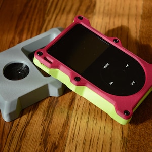 May include: A digital music player with a black circular click wheel and screen, encased in a two-tone magenta and lime green protective frame with black screw accents, positioned next to a separate grey protective case with a circular cutout.