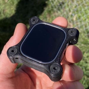 Puede incluir: Un dispositivo electrónico negro con una pantalla cuadrada y un marco texturizado. El marco tiene cuatro cabezas de tornillo en las esquinas. El dispositivo se sostiene en una mano contra un fondo verde borroso.