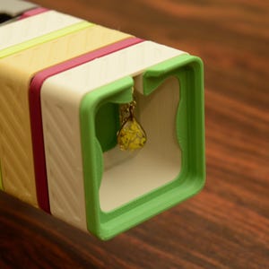 May include: A multi-colored, geometric jewelry box with a gold-toned pendant necklace. The box features a white, green, yellow, and pink design. The pendant is a teardrop shape with yellow flowers encased in clear resin.