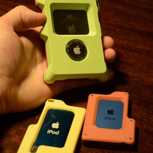 May include: Three portable electronic devices in protective cases. One lime green case holds a black device with an Apple logo and an AirTag. A yellow case holds a blue iPod, and a coral case holds a blue iPod. Each case features a cutout design.