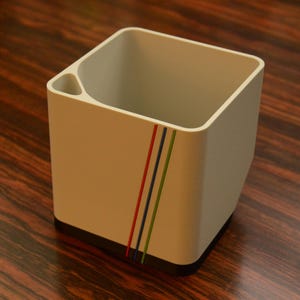 May include: A beige square desk organizer with a black base and a small side compartment. The organizer has three diagonal stripes in red, blue, and green. The organizer is made of a solid material.