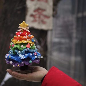 Könnte beinhalten: Ein handgefertigter, kleiner Weihnachtsbaum aus bunten Garnen. Der Baum hat einen Regenbogenfarbverlauf und ist mit kleinen Ornamenten, Perlen und einem gelben Stern verziert. Er steht auf einem Holzsockel und wird in einer Hand gehalten. Der Text "焦美美" ist sichtbar.
