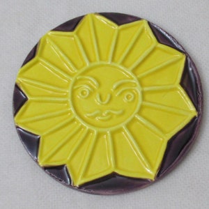 May include: A yellow ceramic sun with a face, surrounded by a dark purple border. The sun is a round coaster.
