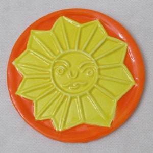 May include: A yellow ceramic sun with a face, surrounded by orange, is a decorative coaster.