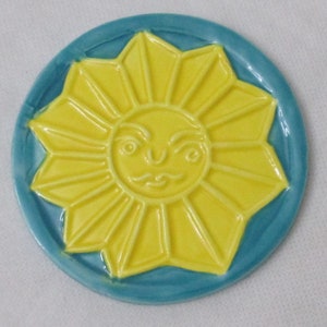 May include: A yellow ceramic sun with a face, surrounded by a blue circle, is a decorative coaster.