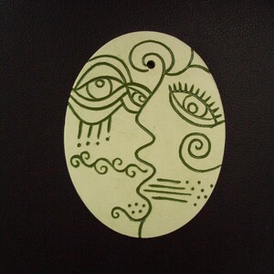 Peut inclure: Une décoration en céramique blanche avec un motif abstrait vert de deux visages avec des yeux, un nez et une bouche. La décoration est de forme ovale.