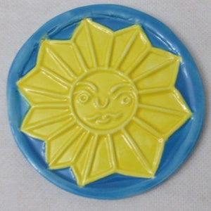 Könnte beinhalten: Eine gelbe Keramik-Sonne mit einem Gesicht, umgeben von einem blauen Kreis.