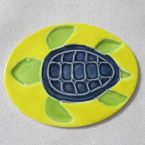 May include: A ceramic tile with a yellow background and a blue and green sea turtle design.