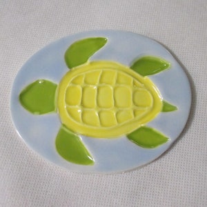 May include: A ceramic tile with a yellow and green turtle design on a light blue background.