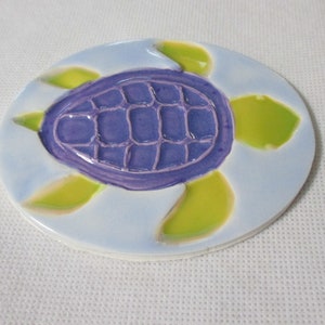 May include: A ceramic coaster featuring a turtle design. The turtle's shell is a grid of purple squares, surrounded by light blue and green accents. The coaster is round and has a glossy finish.